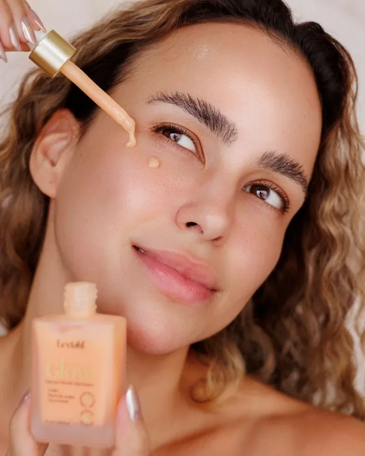 Mulher sorrindo com uma gota do sérum na bochecha, segurando o conta-gotas e o frasco NanoGlow